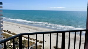 A beach view from a balcony with a railing, sandy beach, ocean, and clear sky.