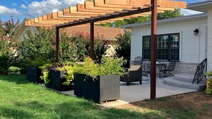 Un'area patio coperta con un pergolato in legno, posti a sedere all'aperto e fioriere con piante fiorite.