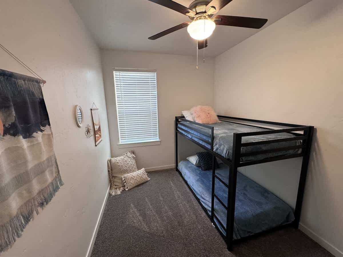 Une petite chambre avec un seul lit, un lit superposé, un ventilateur de plafond, une fenêtre avec des stores et un sol recouvert de moquette.