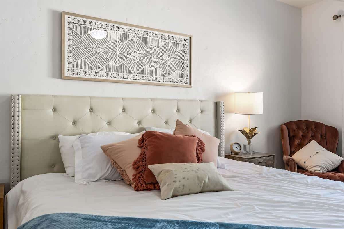 Een net opgemaakt bed met een gestoffeerd hoofdeinde, een decoratief object aan de muur, een bedlampje en een stoel.