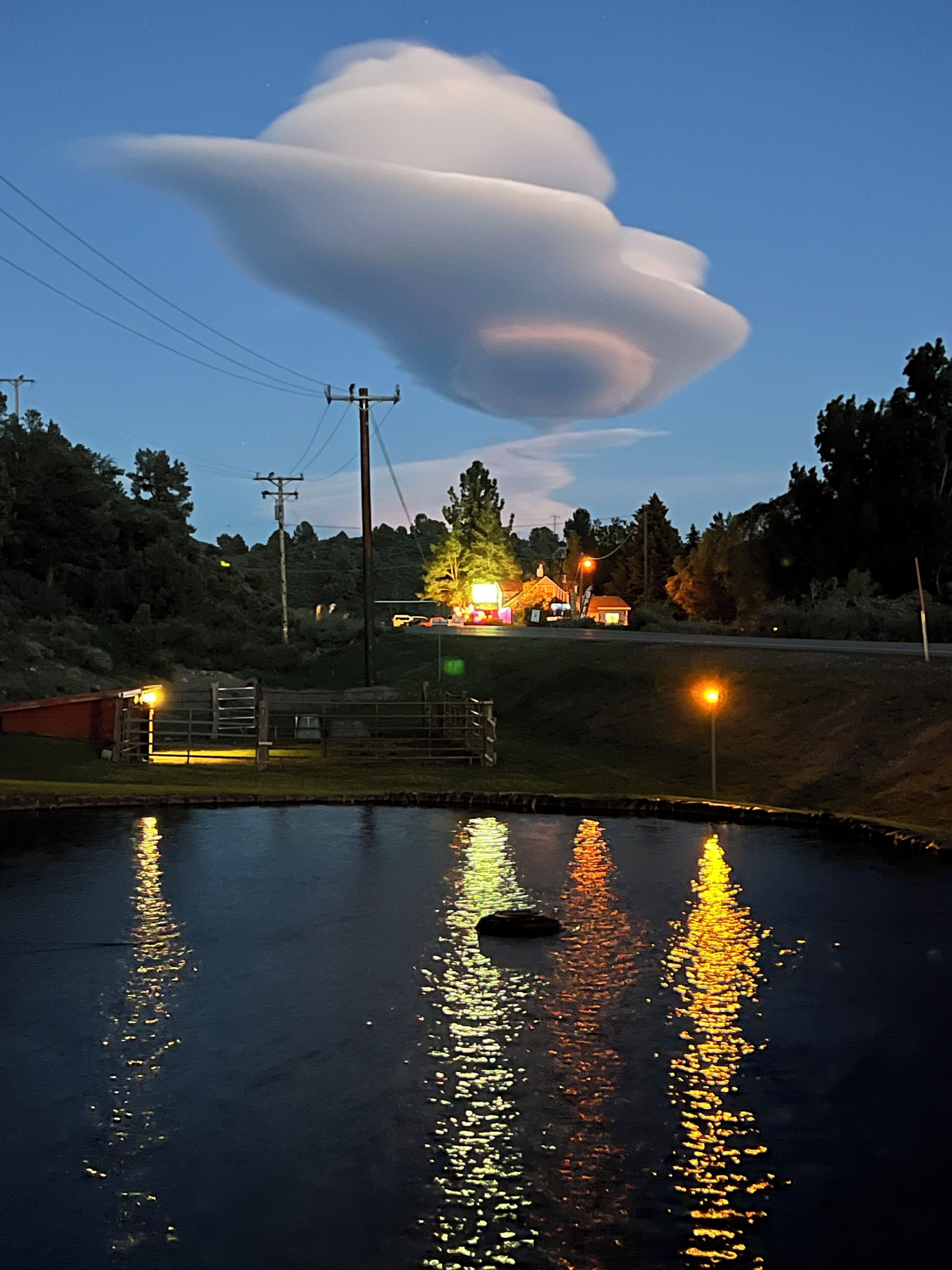 A calm body of water reflecting lights, a building with illuminated windows, and a large cloud formation in the sky.