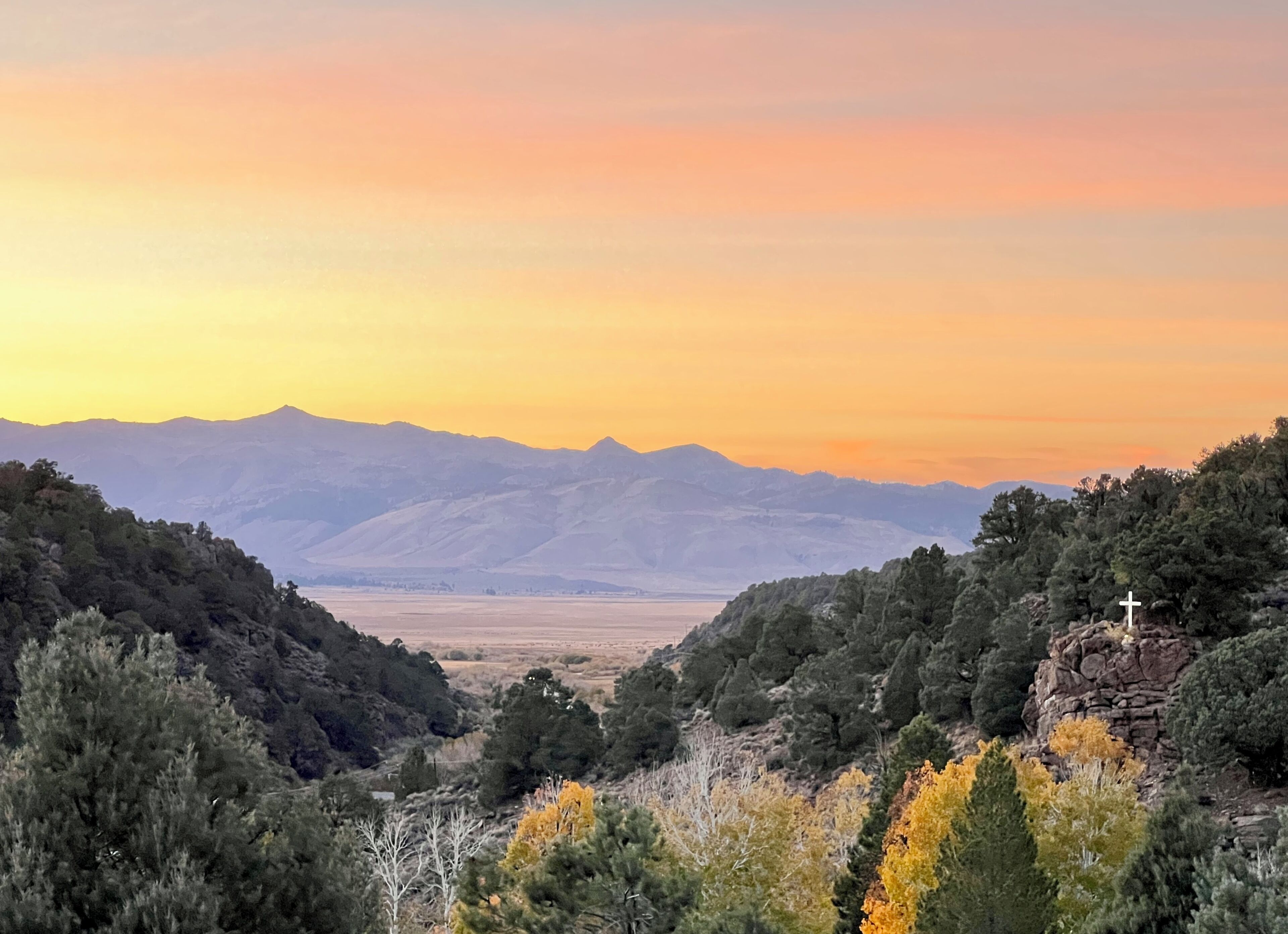 Paisagem montanhosa com uma cruz em afloramento rochoso, rodeada por árvores e um vale.