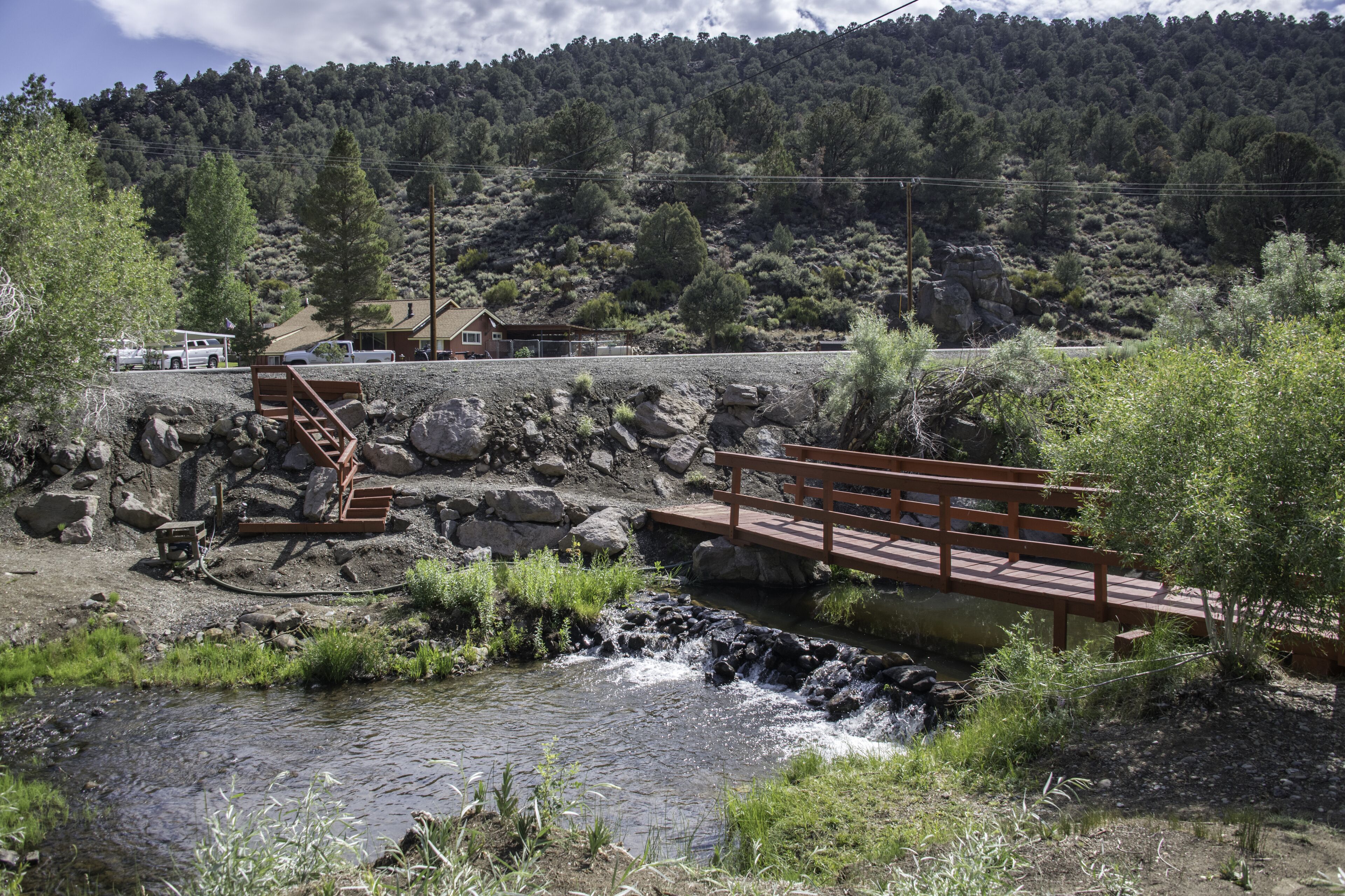 A rustic wooden bridge over a stream, surrounded by greenery and a mountainous backdrop.