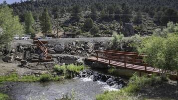 A rustic wooden bridge over a stream, surrounded by greenery and a mountainous backdrop.