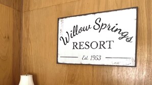 A bedroom with a sign reading "Willow Springs Resort Est. 1953" on the wall, a lamp on a bedside table, and a bed with a deer-themed pillow. - Willow Springs Resort (Bridgeport)