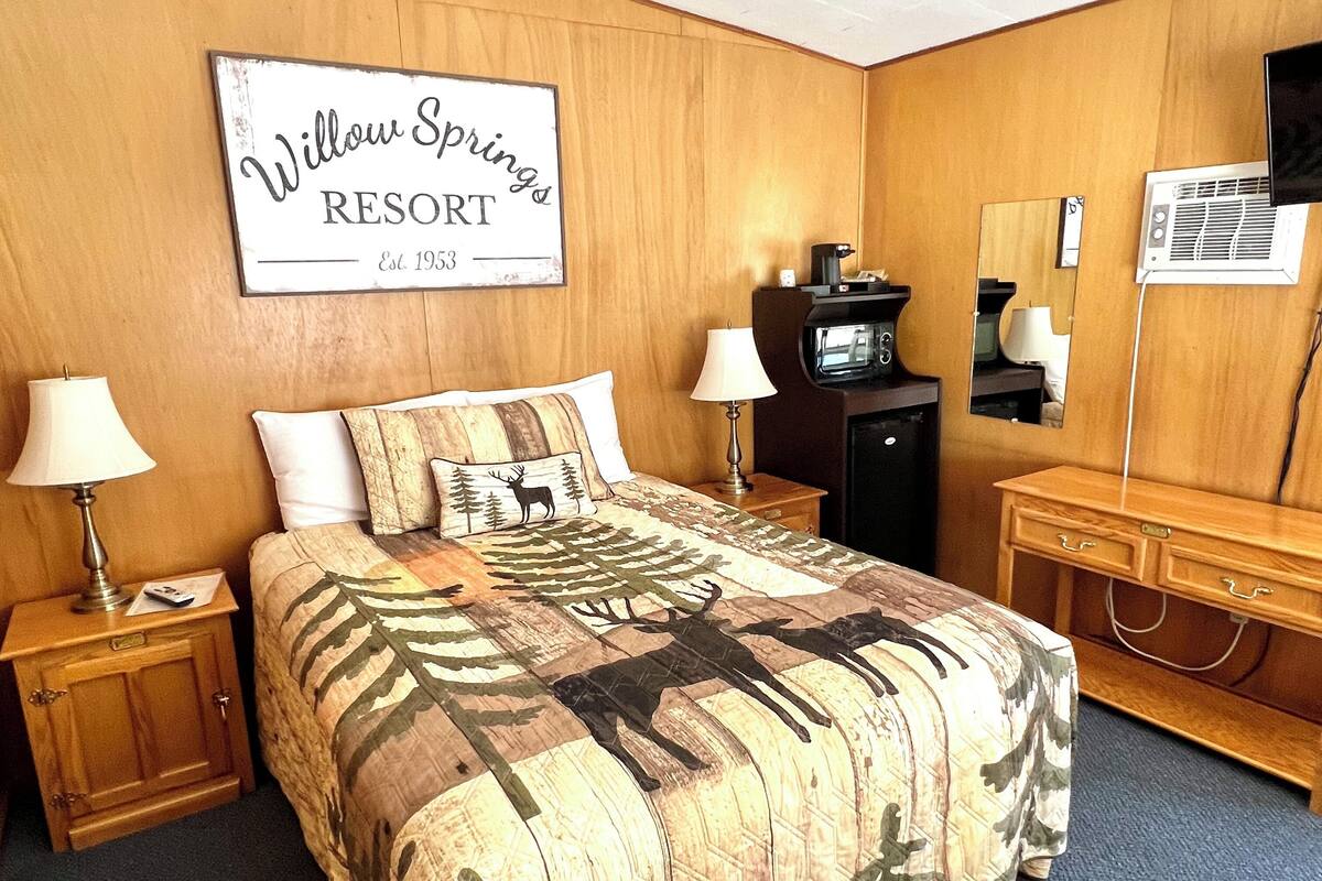 A hotel room with a bed, nightstands, a desk, a mirror, and a sign reading "Willow Springs Resort Est. 1953".