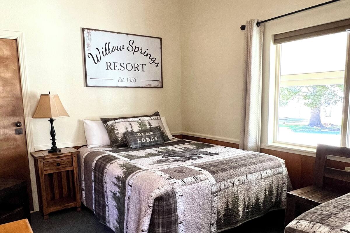 A neatly arranged bedroom with a bed, a nightstand, a lamp, a sign on the wall, and a window with a view.