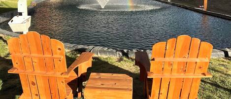 A covered outdoor seating area with Adirondack chairs and a small table, overlooking a pond with a water feature and a rainbow.