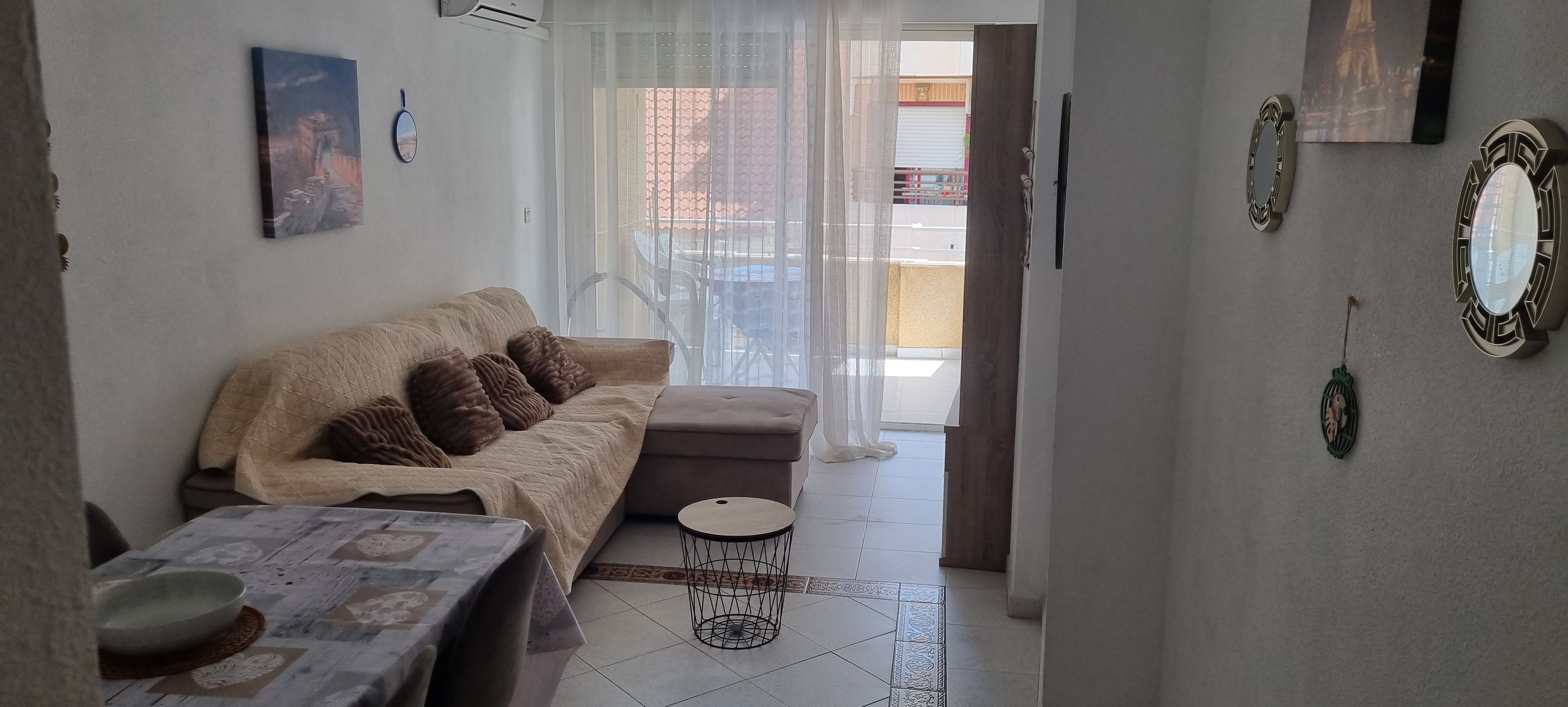 A living room with a sofa, a small round table, and a dining table with chairs. There is a window with sheer curtains, a wall-mounted air conditioner, and a framed picture on the wall.