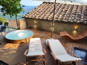 Een houten terras met een jacuzzi, twee lounge fauteuils en een betegeld dak.
