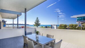 A modern outdoor dining area with a glass table and wicker chairs, overlooking a coastal view with a clear sky. - Beachfront 2 - Steps away from the prestine waters of Mollymook Beach (Mollymook)