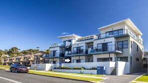 A modern multi-story hotel with balconies, a car parked in front, and a clear blue sky. - Beachfront 2 - Steps away from the prestine waters of Mollymook Beach (Mollymook)