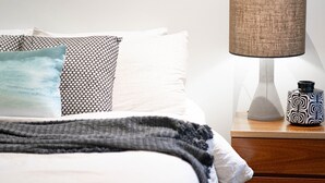 A neatly made bed with a patterned pillow, a plain pillow, and a blue accent pillow. There is a lamp with a textured shade on a wooden nightstand beside the bed. - Beachfront 2 - Steps away from the prestine waters of Mollymook Beach (Mollymook)