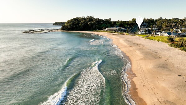 A coastal beach with a clear view of the ocean, a sandy shore, and a residential area with houses in the background.