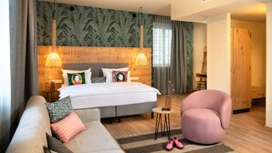 A modern hotel room with a large bed, two bedside lamps, a wooden headboard, a grey sofa, a pink armchair, and a small round table with shoes. - Hotel Das Regensburg (Regensburg)