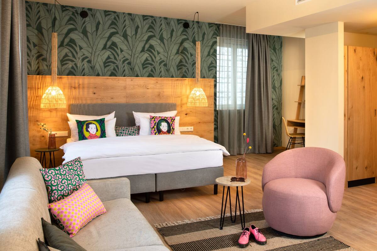 Una habitación de hotel moderna con una cama grande, dos lámparas de noche, un cabecero de madera, un sofá gris, un sillón rosa y una mesita redonda con zapatos.