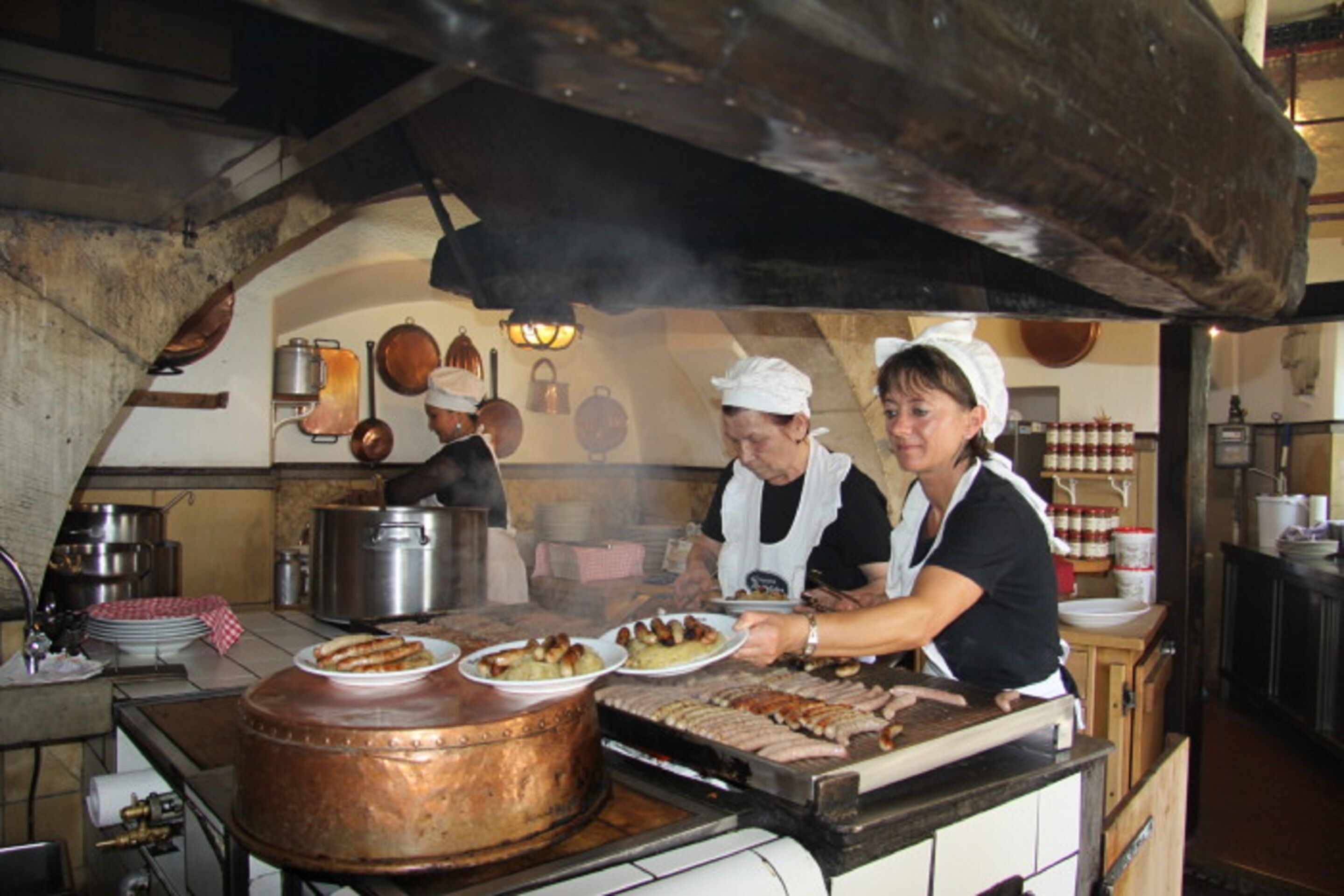 Una cocina con una olla de cobre grande, una parrilla y dos cocineros preparando comida.
