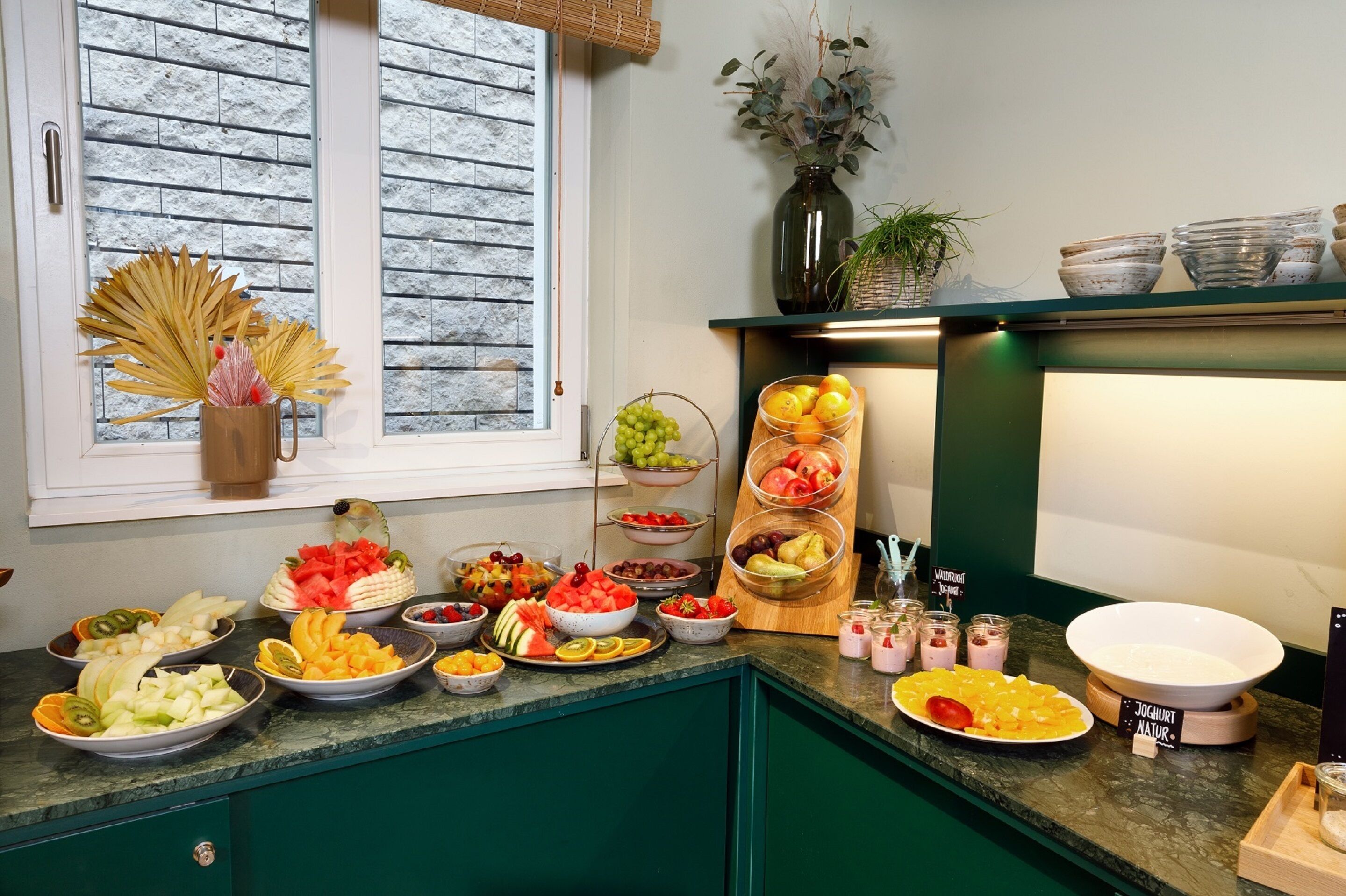 Ein Buffet mit verschiedenen Früchten und Gemüsesorten, eine Vase mit Trockenpflanzen und ein Fenster mit Blick nach draußen.