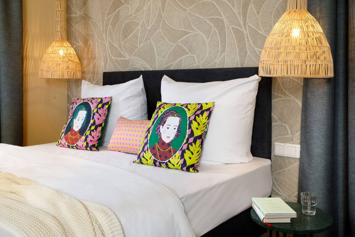 Una cama bien tendida con sábanas blancas, dos almohadones decorativos, una mesita de noche con un libro y un vaso de agua, y dos lámparas tejidas.