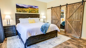 A bedroom with a large bed, wooden headboard, two bedside tables with lamps, a large wooden sliding door, and a painting on the wall.