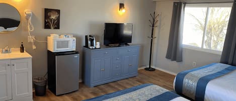 A hotel room with a bed, a desk with a computer, a microwave, a coffee maker, a TV, and a window with curtains.