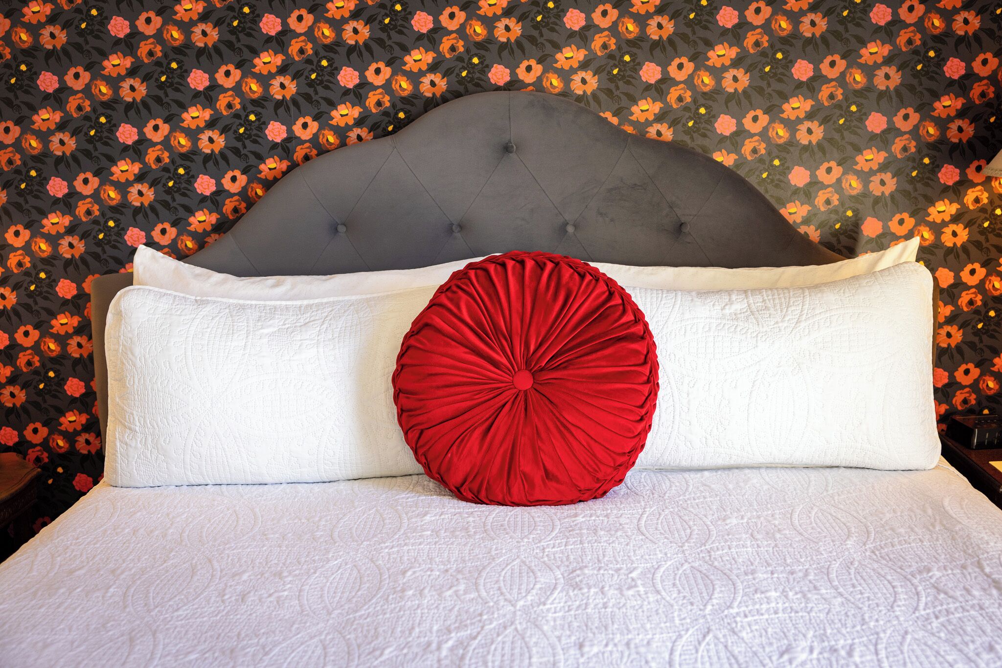 Una cama con cabecera gris tapizada, ropa de cama blanca y una almohada decorativa roja.