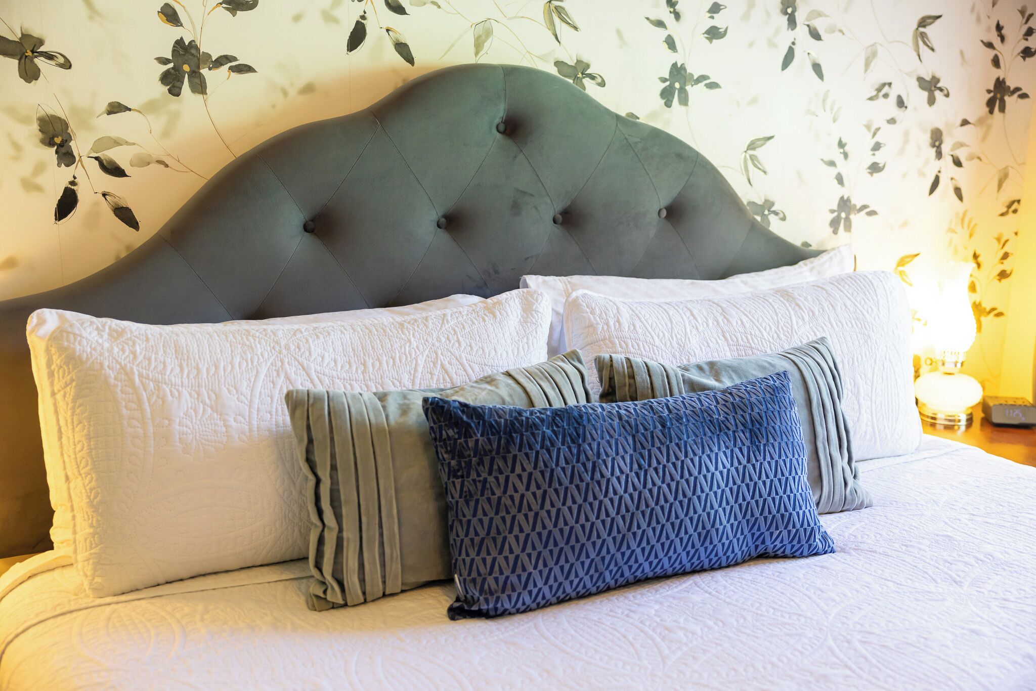 A bed with a tufted headboard, white and patterned pillows, and a floral wallpaper background.
