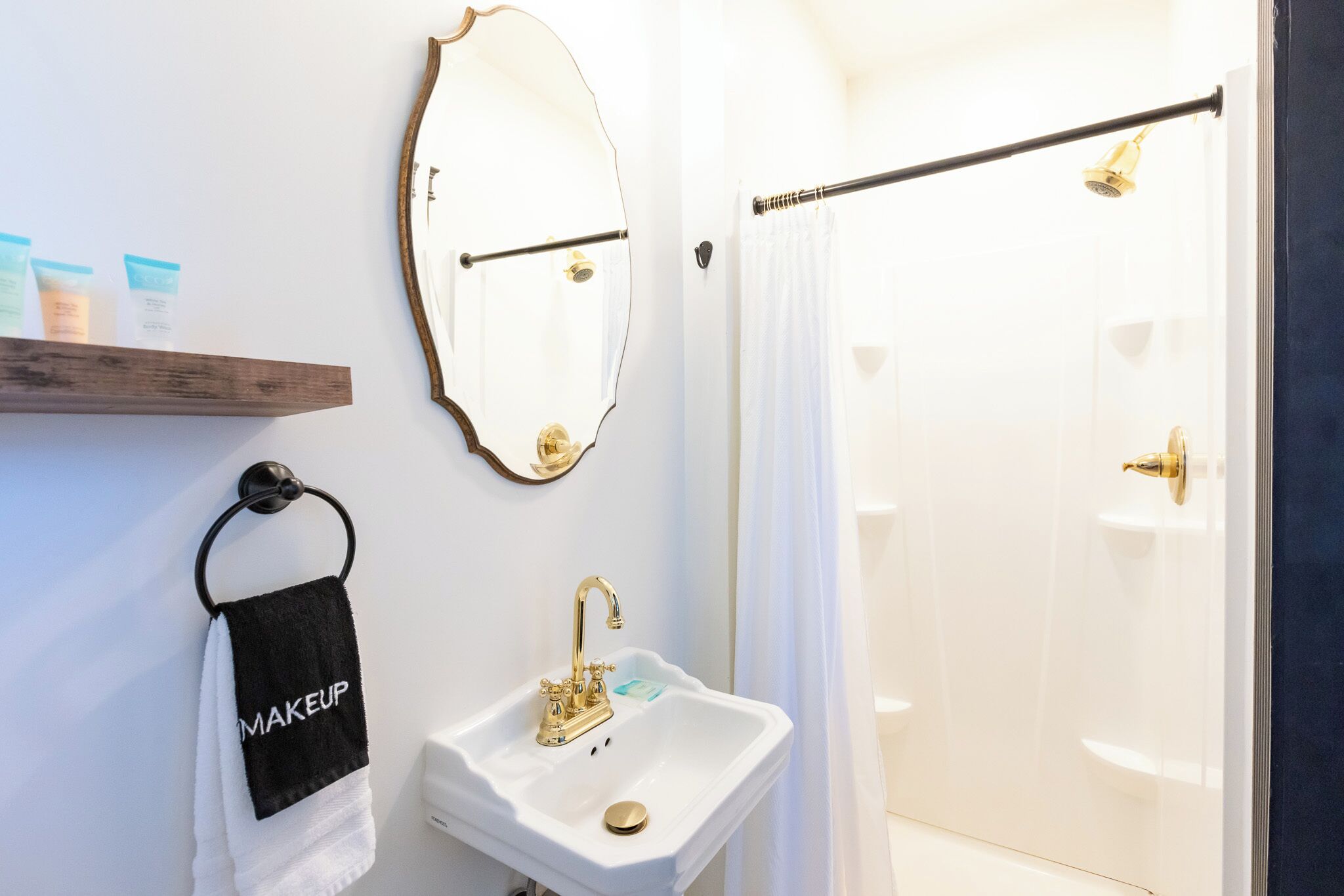Un baño con un lavamanos blanco, grifo dorado y un espejo con marco dorado. Se observa un perchero con una toalla negra que dice 'MAKEUP'.