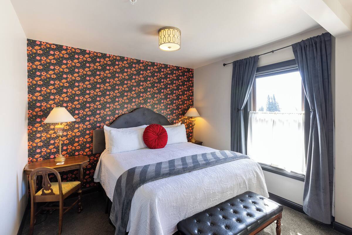 A hotel room with a bed, bedside lamps, a small table with a chair, a bench, and a window with curtains.