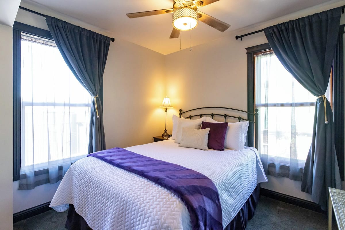 A hotel room with a bed, a ceiling fan, a nightstand, and a window with curtains.