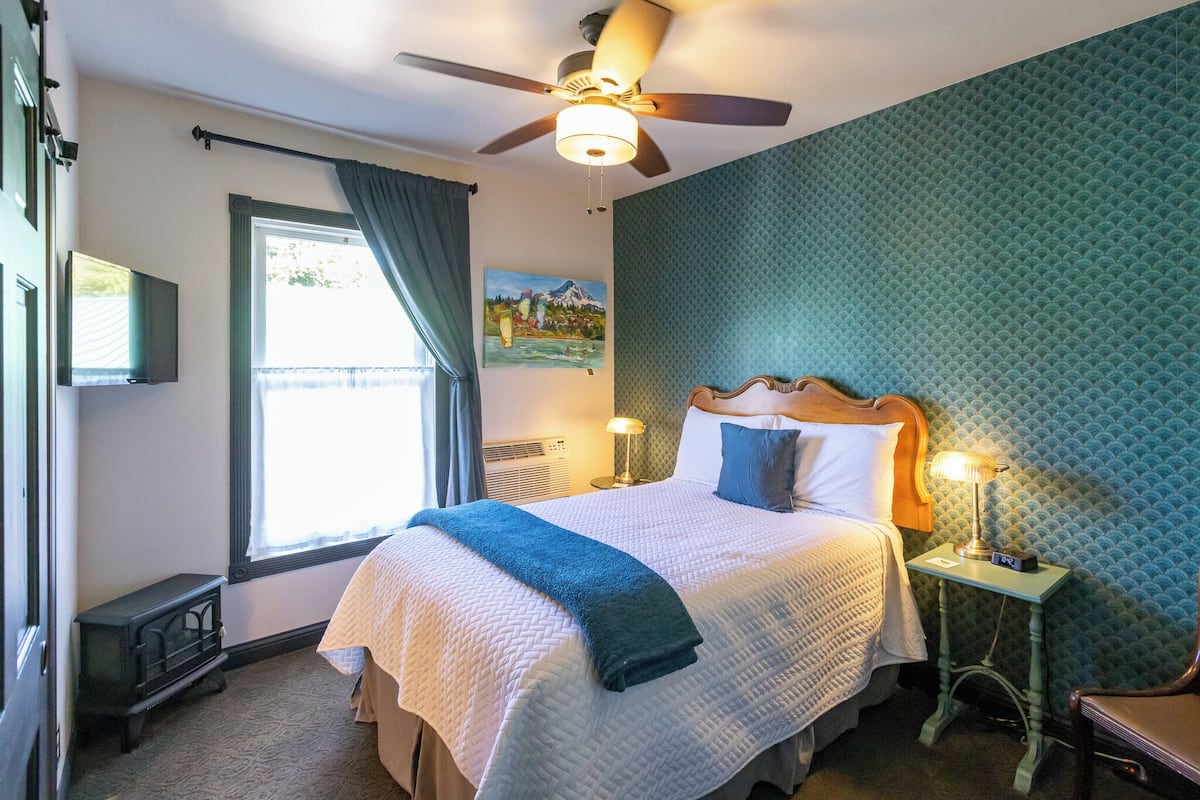 A neatly made bed with a blue blanket, a ceiling fan, a bedside table with a lamp, and a painting on the wall.
