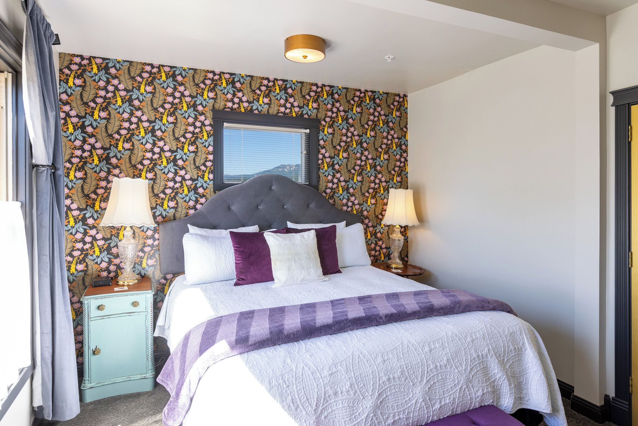 A bedroom with a floral wallpaper, a tufted headboard, two bedside tables with lamps, and a window with blinds.