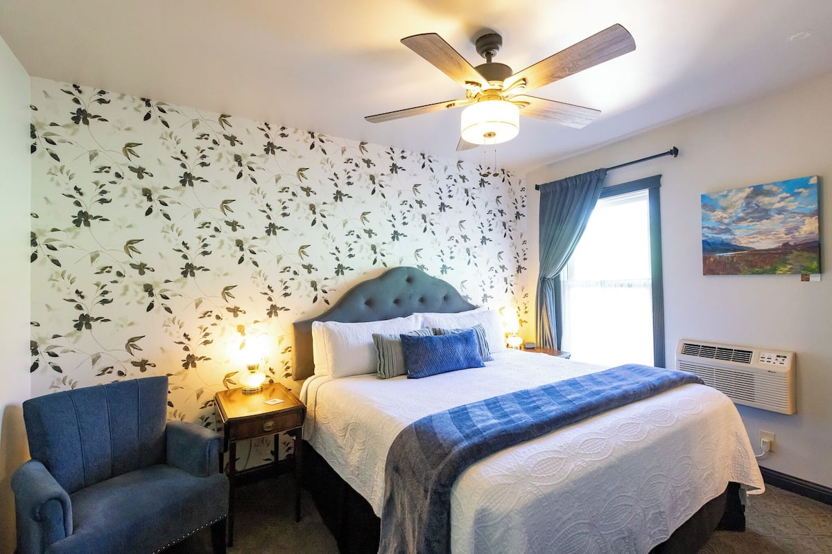 A bedroom with a bed, a chair, a ceiling fan, a window with curtains, and a wall-mounted air conditioner.