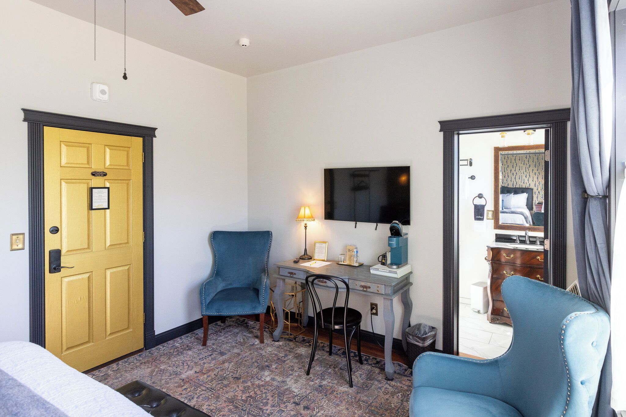 A room with a yellow door, a desk with a lamp, two chairs, a bed, and a television on the wall.