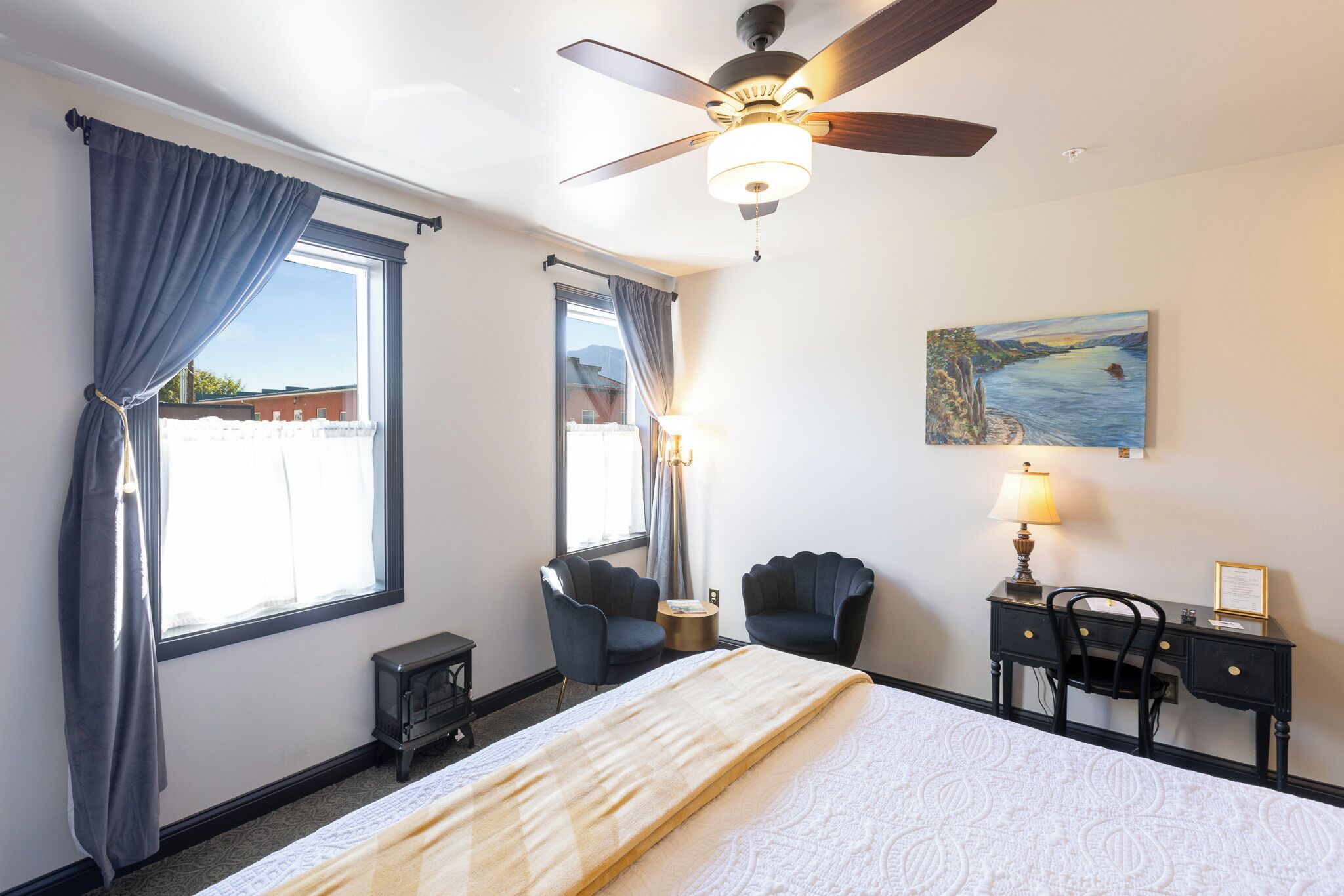 A hotel room with a bed, two chairs, a desk, a ceiling fan, and a painting on the wall.