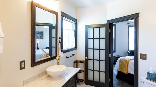 A bathroom with a white marble sink, a large mirror, and a door leading to a bedroom. - Hotel Stevenson (Stevenson)