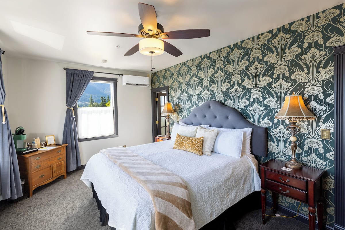 A bedroom with a large bed, a ceiling fan, a nightstand with a lamp, a dresser, and a window with curtains.