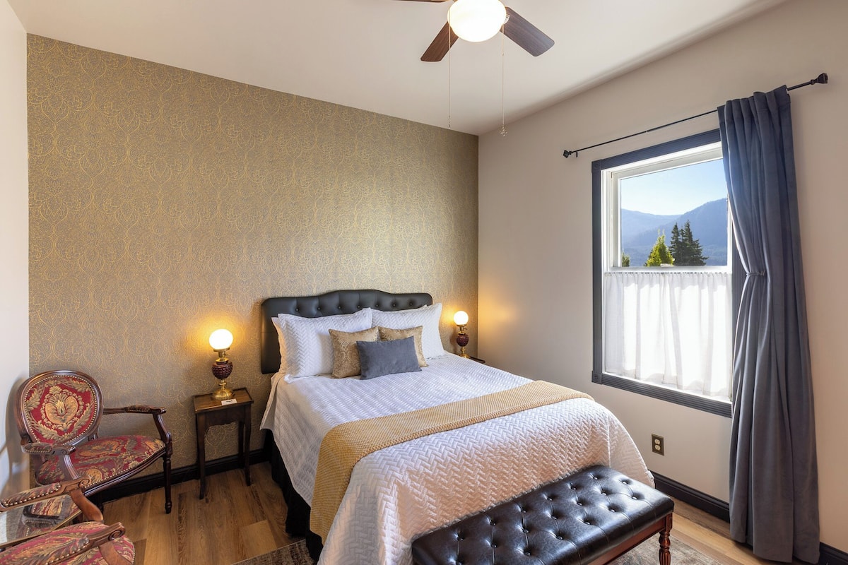 A bedroom with a bed, a chair, a bedside table, a lamp, a window with curtains, and a view of mountains.