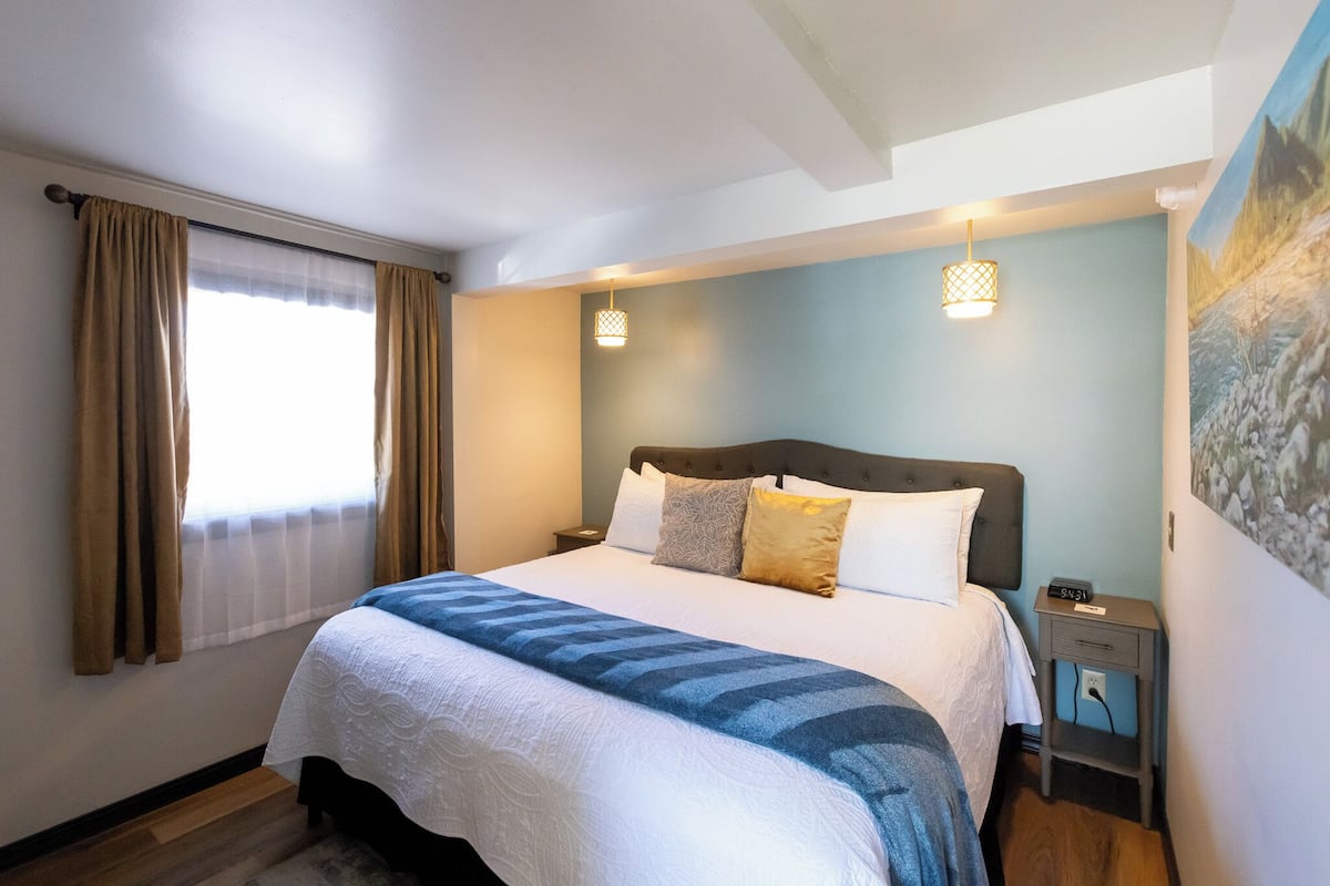 A neatly made bed with a blue and white bedspread, a bedside table with a lamp, and a painting on the wall.