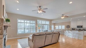 Un salón espacioso con un amplio sofá seccional color beige, una chimenea y una zona de cocina con armarios blancos y una mesa de comedor.