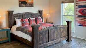 A bedroom with a wooden bed, a painting of a landscape, a window with a view, and a rug.