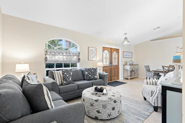 A living room with a grey sofa, a round coffee table, and striped chairs. There is a window with a view of greenery, a door with glass panels, and a chandelier.