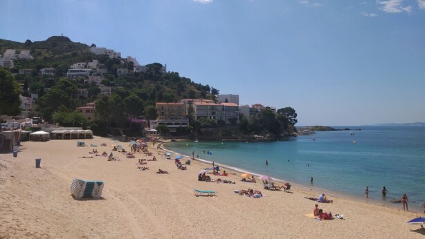 Una playa con gente tomando el sol y nadando, rodeada de edificios y colinas.