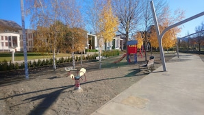Ein Spielplatz mit Rutsche und Schaukeln, umgeben von Bäumen mit Herbstlaub, und ein Gebäude im Hintergrund.