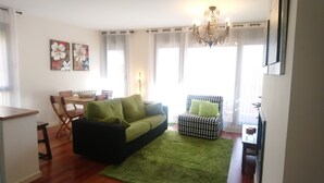 Ein Wohnzimmer mit einem grünen Sofa, einem gemusterten Sessel, einem Esstisch mit Stühlen und einer Chandelüster.