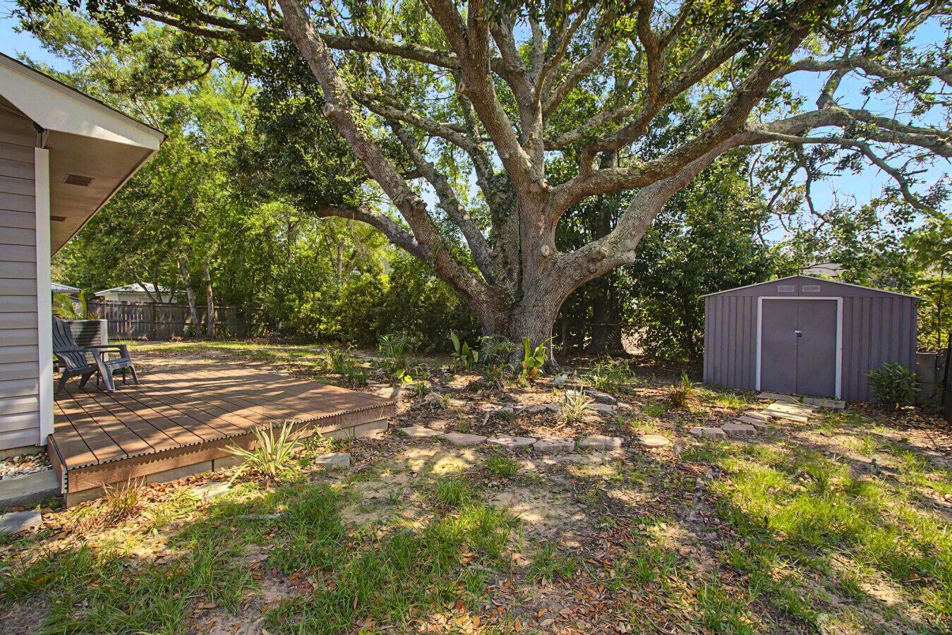 A backyard with a large tree, a patio with chairs, a shed, and a house.