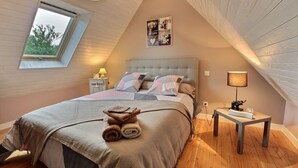 Une chambre avec un plafond en pente, un grand lit avec une tête de lit, une table de chevet avec une lampe et une fenêtre donnant sur des arbres.