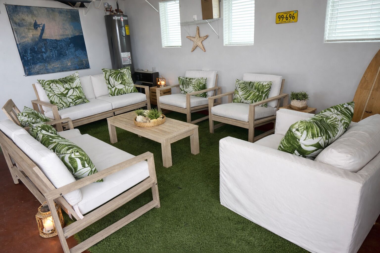 Un salon doté d’un sol recouvert d’un gazon artificiel vert, de canapés blancs ornés de coussins à motifs de feuilles vertes, d’une table basse en bois et d’une décoration en forme d’étoile de mer accrochée au mur.