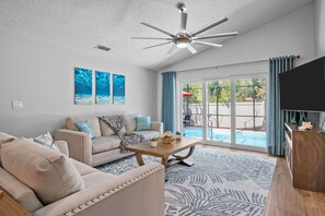 Ein Wohnzimmer mit Deckenventilator, einer Couch, einem Couchtisch und Blick auf einen Swimmingpool durch große Glasüren.