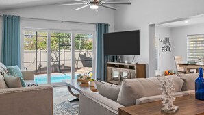 Una sala moderna con un televisor de pantalla plana, una mesa de centro de madera, un sofá color beige y un ventanal con vistas a una piscina.